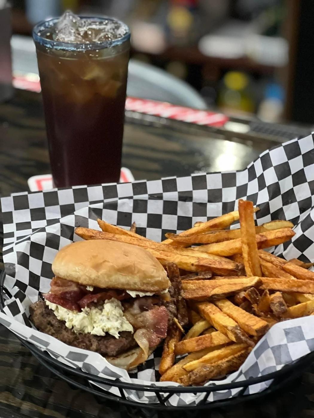 A bacon burger in a basket with hand cut fries and an iced tea, on the dark wood bar top